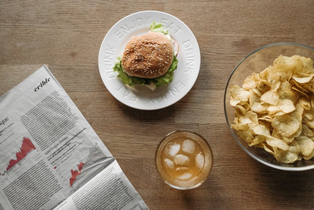 Overhead shot of a burger, chips, and drink on a wooden table with a newspaper.